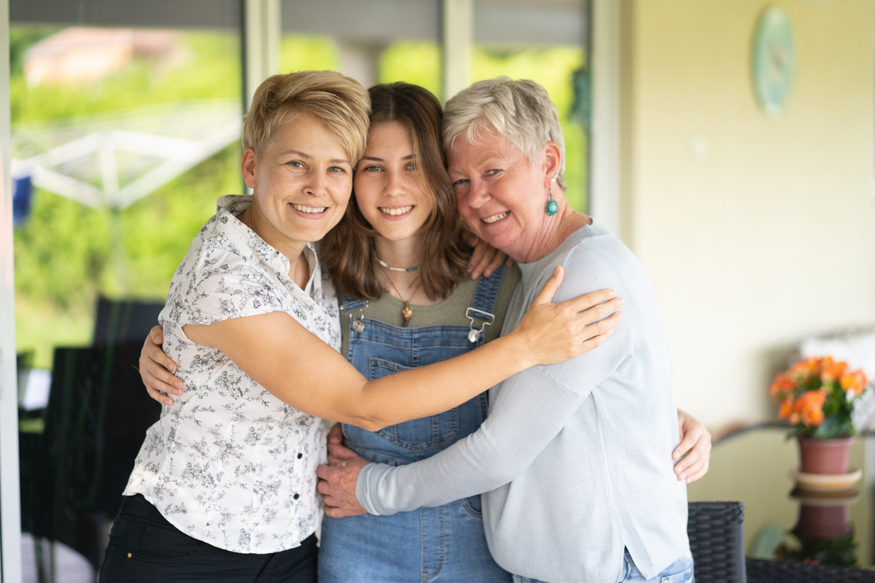 A grey-haired grandmother, with her blonde-haired daughter and brown-haired granddaughter are all hugging and looking at the camera