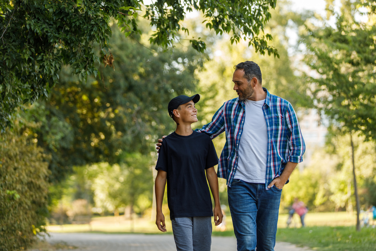 Copy space shot of contented mid adult man embracing his teenage son while they are taking a relaxing walk at the park. They are looking and smiling at each other, having a bonding moment.