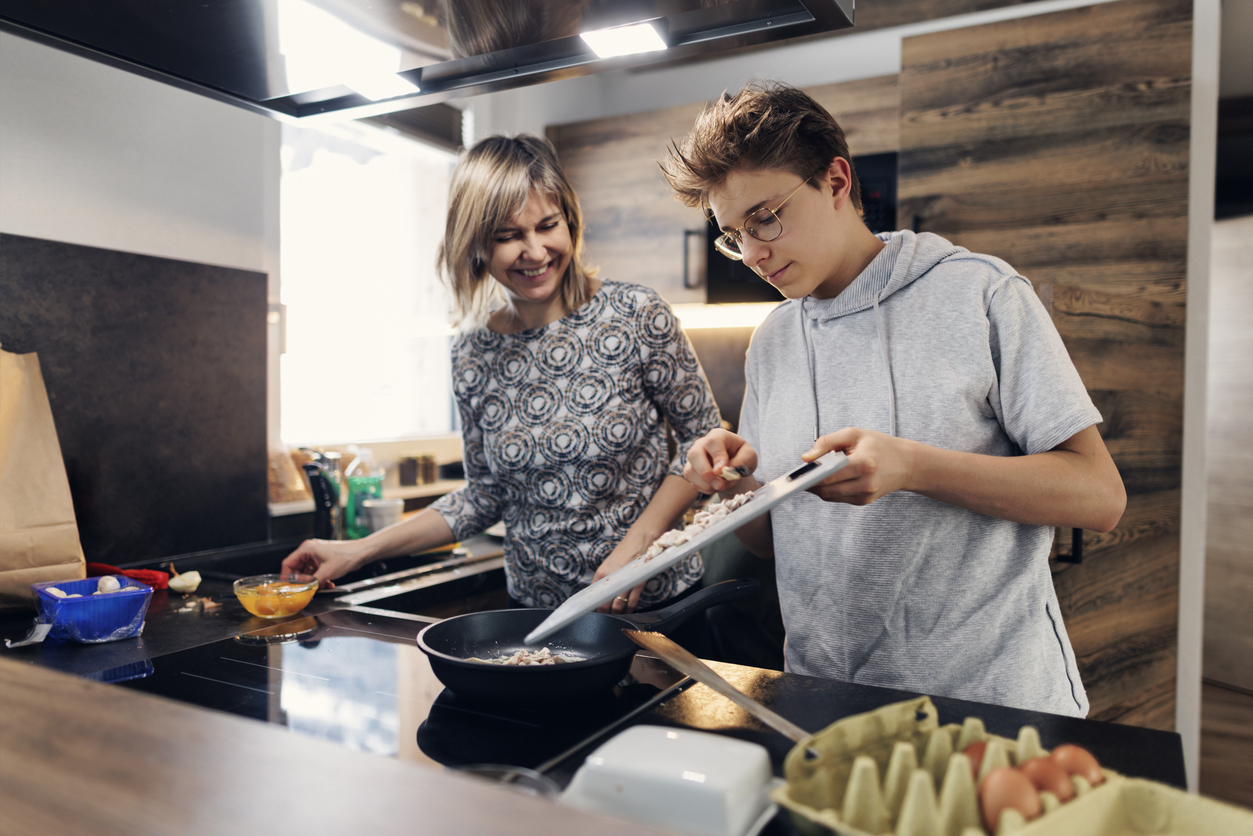 Mother and teenage son are preparing breakfast together. They are making the scrambled eggs. The boy is adding the chopped mushrooms to the frying pan.